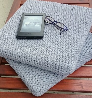 Zwei hellgraue gehäkelte Kissen mit einem Lesegerät und einer Lesebrille auf einer Holzoberfläche.