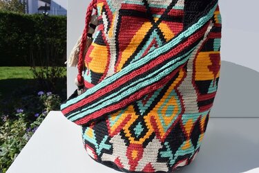 Gehäkelte Tasche mit geometrischem Ethno-Muster in Rot, Gelb, Türkis und Schwarz auf weißer Fläche im Freien.