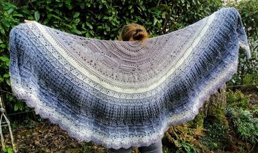 Person hält großes halbrundes gehäkeltes Tuch mit Farbverlauf in Grau, Flieder und Creme, gewellte Spitzenkante sichtbar