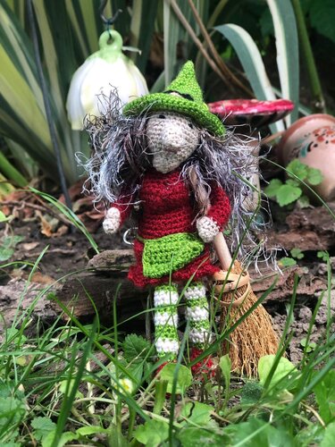 Gehäkelte Hexenpuppe mit grünem Hut, rotem Kleid und Besen im Gras