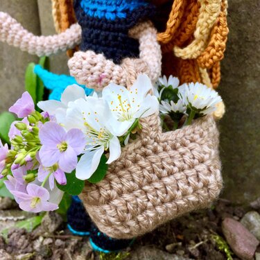 Nahaufnahme einer gehäkelten Figur mit kleinem beigen Korb voller weißer und violetter Frühlingsblumen