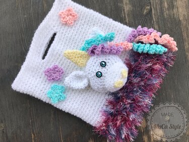 Weiße gehäkelte Tasche mit Einhornkopf, pastellfarbener Mähne, Blumenapplikationen und buntem Fransenrand auf dunklem Holz