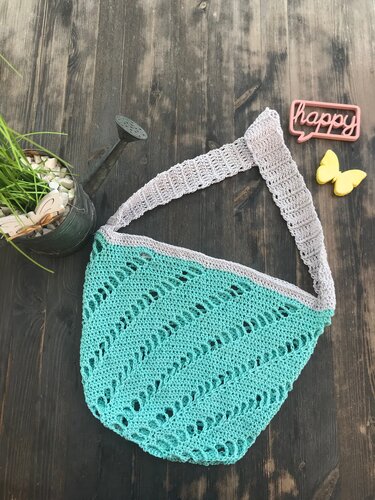 Gehäkelte türkise Umhängetasche mit hellgrauem Träger und diagonalem Lochmuster auf dunklem Holzhintergrund.