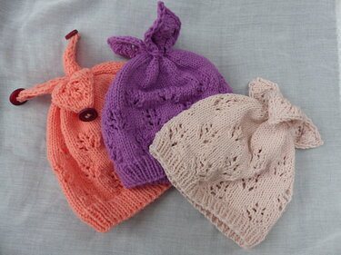 Drei handgestrickte Kindermützen in Apricot, Lila und Rosa mit Lochmuster und dekorativen Details