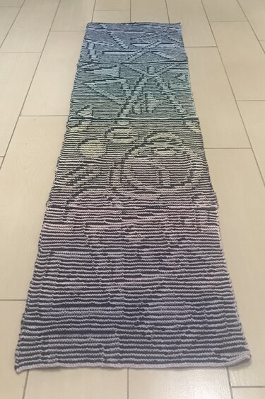 Langer gestrickter Schal mit Farbverlauf und geometrischen Mustern auf hellem Fliesenboden