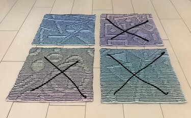 Vier gestrickte Maschenproben mit Farbverläufen in Blau, Lila, Grau und Türkis, drei davon mit schwarzem X.