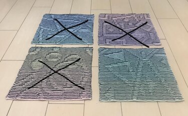 Vier gestrickte Musterproben mit Farbverläufen und Streifen auf hellem Fliesenboden, drei mit schwarzem X markiert