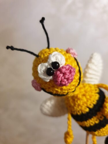 Nahaufnahme einer gelb‑schwarzen gehäkelten Amigurumi‑Biene mit weißen Flügeln, rosa Wangen und schwarzen Perlenaugen