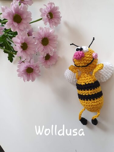 Gehäkelte Amigurumi-Biene in Gelb und Schwarz mit weißen Flügeln neben rosa Blüten
