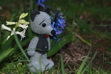 Gehäkeltes graues Amigurumi-Tier mit spitzen Ohren im Gras neben Blumen