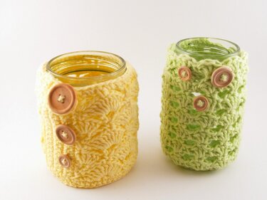 Zwei gehäkelte Glasüberzüge in Gelb und Hellgrün mit Holzknöpfen auf hellem Hintergrund