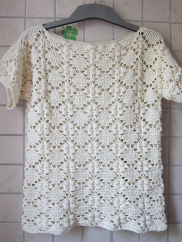 Cremefarbenes gehäkeltes Damen-Shirt mit kurzem Ärmel und dekorativem Loch- und Noppenmuster, an einem Kleiderbügel hängend.