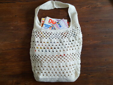 Weiße gehäkelte Netz-Tasche mit breitem Henkel, gefüllt mit Zeitschriften, auf Holztisch