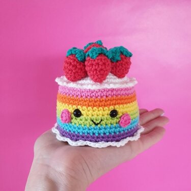 Hand holding a small rainbow crochet cake with strawberries and a smiling face against a pink background