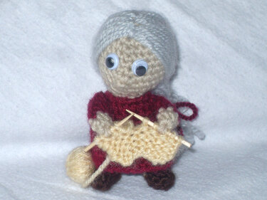 Gehäkelte Amigurumi-Puppe einer strickenden Großmutter mit grauem Haar, rotem Kleid, Stricknadeln und Garn.