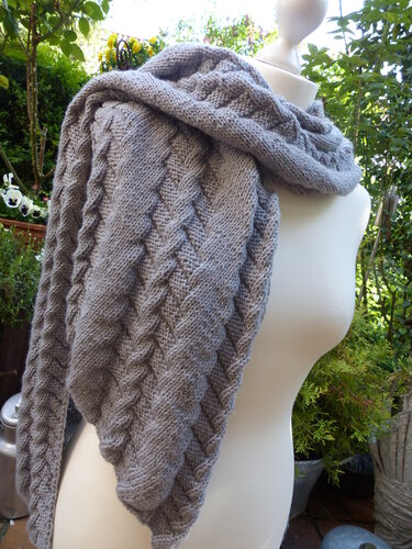 Gestrickter grauer Schal mit Zopfmuster auf einer Schaufensterpuppe, im Hintergrund Pflanzen