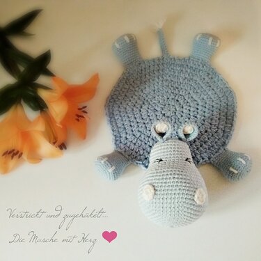 Hellblaues gehäkeltes Nilpferd‑Schmusetuch auf hellem Untergrund neben orangefarbenen Lilien.