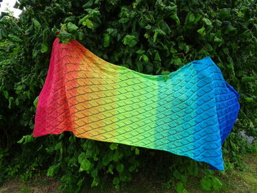 Bunter Strickschal mit Regenbogen-Farbverlauf und Rauten-Ajourmuster vor grünem Laub.