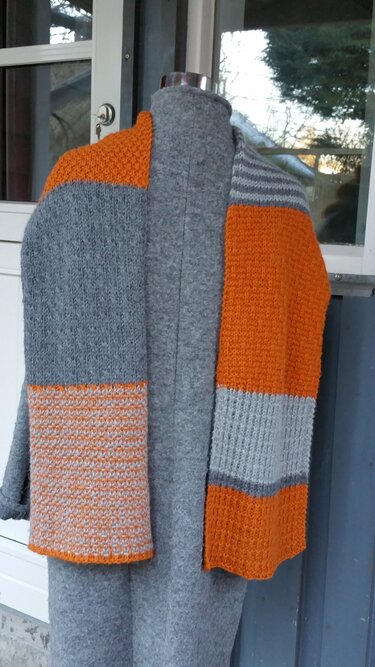 Gestrickter Schal in Grau und Orange mit Blockstreifen über einer Schneiderpuppe drapiert, vor einem Fensterhintergrund