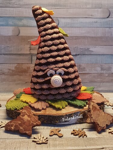 Gehäkelte braun-beige Baumkreatur mit großen Augen und kleiner Nase auf Holzscheibe, umgeben von gehäkelten Herbstblättern in Grün, Gelb und Orange.