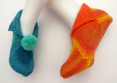 Zwei gestrickte Hausschuhe an Schaufensterbeinen: links petrol mit Bommel, rechts orange-gelb mit umgeschlagener Krempe