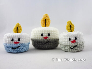 Drei runde gehäkelte Teelicht-Figuren mit Gesichtern und gelben Flammen in Weiß, Hellblau, Dunkelgrau und Hellgrau vor weißem Hintergrund.