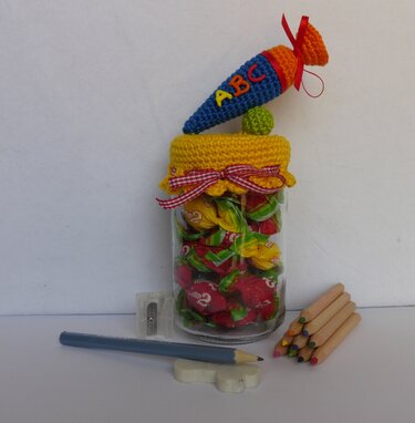 Glas mit gehäkeltem gelben Deckel und Mini-Schultüte, gefüllt mit bunten Bonbons; Buntstifte, Radiergummi und Spitzer daneben