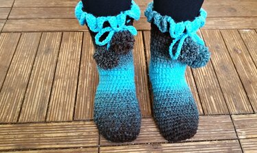 Füße in türkisen bis grauen gehäkelten Haussocken mit Rüsche und Bommel auf Holzterrasse