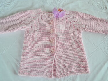 Rosa gestrickte Babyjacke mit langen Ärmeln, Knopfleiste und Blättermuster an den Schultern. Kleine lila Stoffblüte am Kragen.