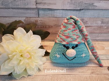 Gehäkelter Amigurumi-Wichtel in Türkis mit mehrfarbiger Zipfelmütze neben weißer Dekoblume