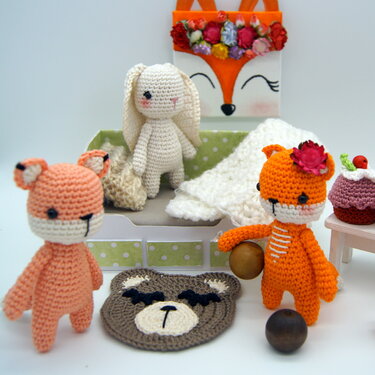 Gehäkelte Amigurumi-Figuren: Fuchs, Hase und Bär mit Mini-Bett und Decke, Bärenteppich und Häkel-Cupcake auf kleinem Tisch.