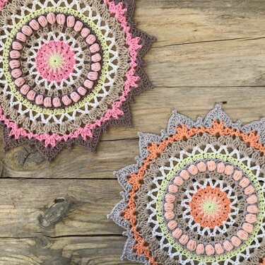 Zwei runde, gehäkelte Mandalas in Pastellfarben mit gezacktem Rand auf Holzuntergrund.