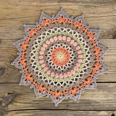 Round crocheted mandala doily with peach, beige, cream and pale green rings on a wooden plank background
