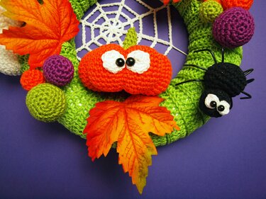 Grün umhäkelter Halloween-Türkranz mit gehäkeltem Kürbis, schwarzer Spinne, weißem Spinnennetz, bunten Häkelkugeln und Herbstblättern vor violettem Hintergrund.