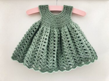 Sage green crocheted baby dress with openwork stitch and scalloped hem on a pink hanger.
