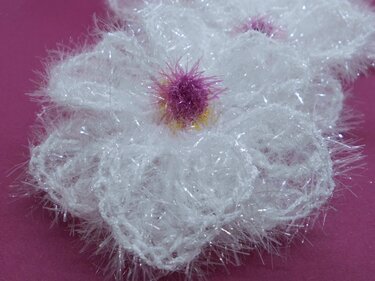 Weiße flauschige Häkelblume aus Fransengarn mit pink-gelber Mitte auf pinkem Untergrund.