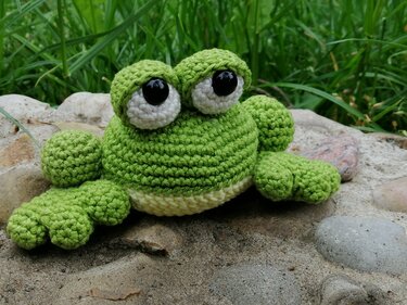 Gehäkelter grüner Amigurumi-Frosch mit großen Augen auf Steinen und Sand vor Gras