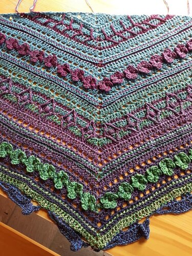 Close-up of a crocheted triangle shawl in blue, green, and purple showing openwork lace, textured stitch rows, and a scalloped edge.