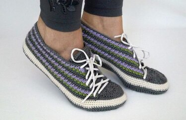 Pair of women's crocheted sneaker-style slippers in grey, purple and green stripes with white laces and white sole edge.