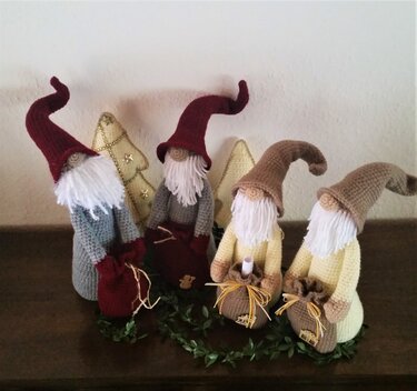 Vier gehäkelte Wichtel mit weißen Bärten und roten sowie beigefarbenen Zipfelmützen, jeweils mit kleinem Säckchen vor goldener Weihnachtsdekoration und grüner Girlande.