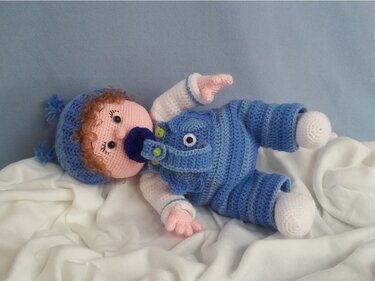 Gehäkelte Babypuppe mit hellblau gestreiftem Strampler, Mütze und Schnuller auf weißer Unterlage.