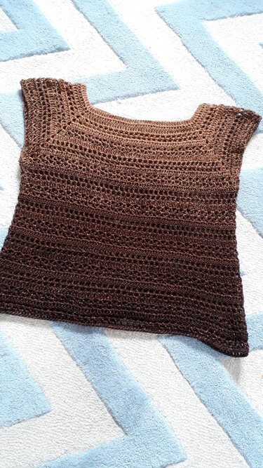 Ärmelloses gehäkeltes Damen-Shirt in Braun mit Ombré-Effekt und Lochmuster auf hellblau-weiß gemustertem Untergrund liegend.