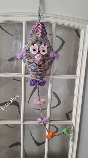 Gehäkelte diamantförmige Amigurumi-Dekoration in buntem Garn mit Augen, Lächeln und Schleifen, an einer Tür hängend.