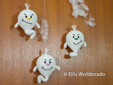 Drei weiße gehäkelte Amigurumi-Geister mit verschiedenen Nasenfarben an Schnüren vor Holz