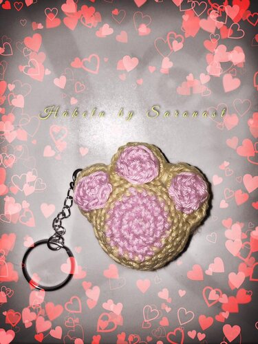 Gehäkelter Pfoten-Schlüsselanhänger in Beige mit rosa Pfotenballen und silbernem Schlüsselring auf gemustertem Hintergrund