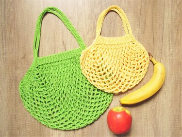 Zwei gehäkelte Netztaschen, eine grün und eine gelb, auf Holzuntergrund; Banane und Apfel als Größenvergleich