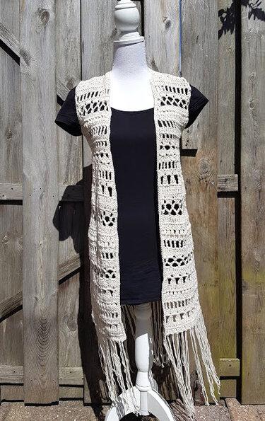 Long sleeveless beige crochet vest with openwork panels and long fringe on a mannequin in front of a wooden fence.
