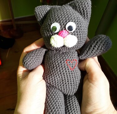 Handgehaltene graue gehäkelte Amigurumi‑Katze mit weißen Schnauzenpolstern, rosa Nase und gesticktem roten Herz
