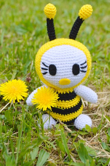 Gehäkelte Amigurumi-Biene in Gelb-Schwarz mit Fühlern, sitzend im Gras neben Löwenzahn