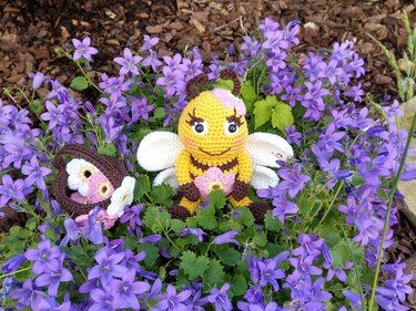 Gelb-braune Amigurumi-Biene neben einem gehäkelten Korb, umgeben von lila Blüten